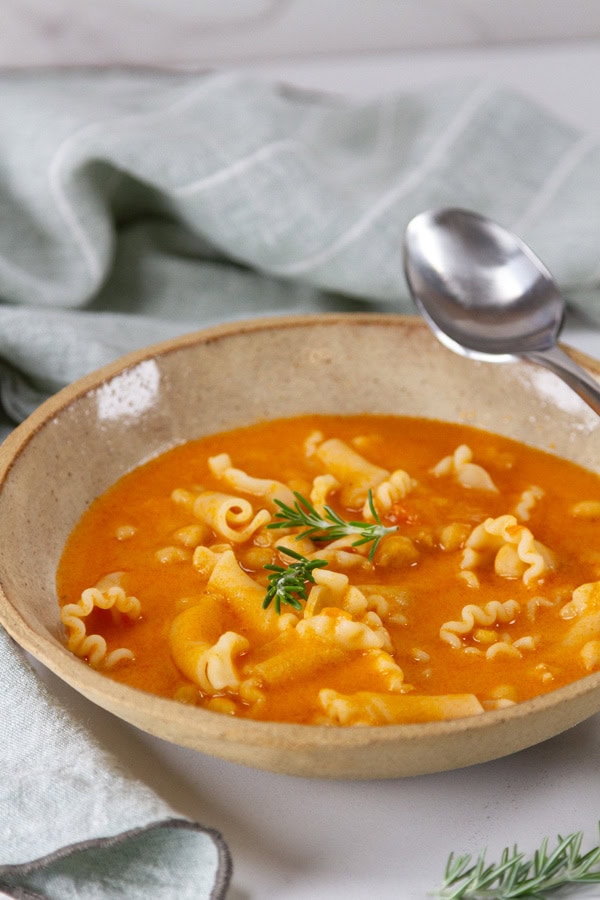 a bowl of tomato noodle chickpea soup with a spoon on the right.