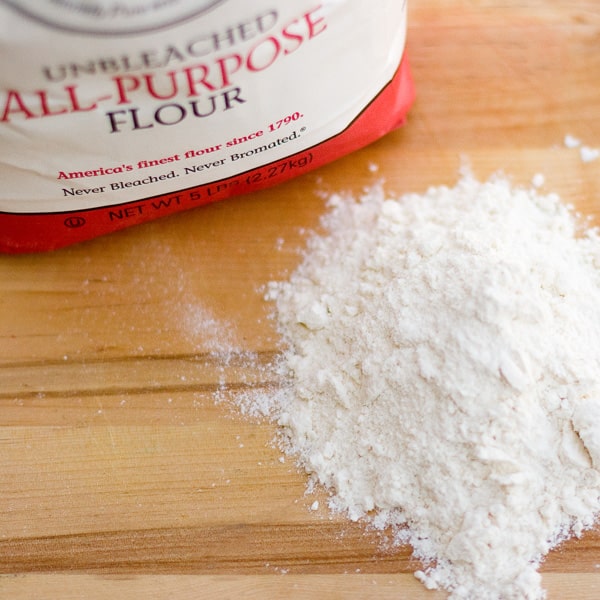 a pile of all purpose flour next to the bag on a wooden background.