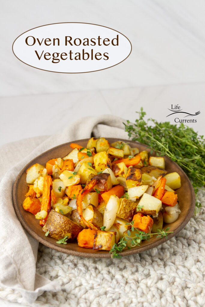 A brown bowl filled with veggies on a white placemat with a cloth napkin, fresh herbs around, title on image: Oven Roasted Vegetables