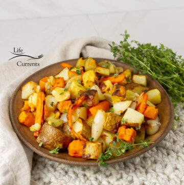square crop of a bowl full of roasted vegetables with fresh herbs around.