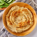 square crop of a bowl of hummus on a cloth with veggies next to it.