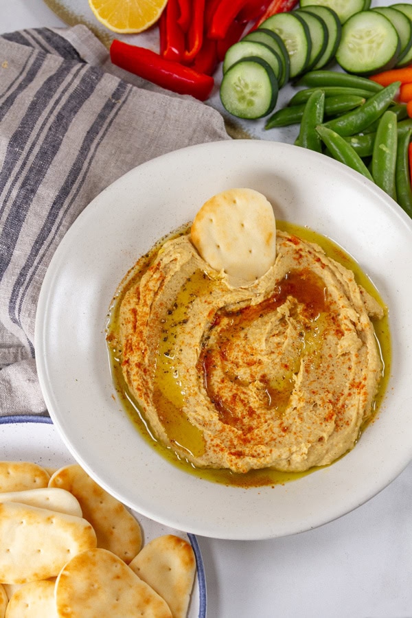 hummus in a white bowl with a naan and some veggies around the bowl.