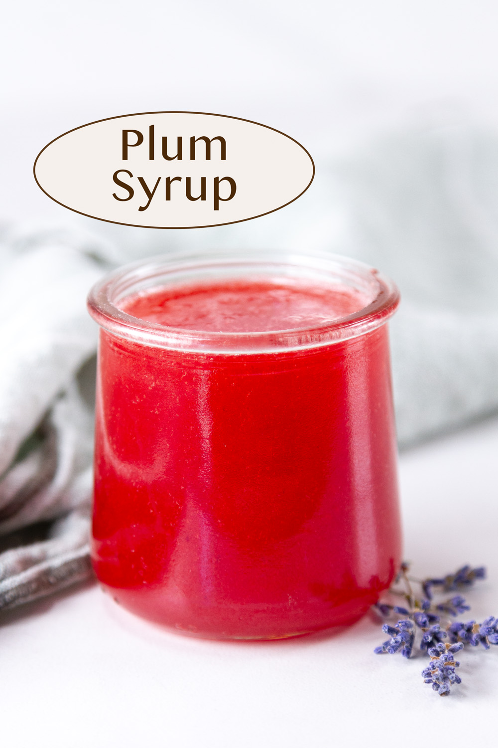 plum syrup in a small glass jar in front of a green cloth napkin and some lavender flowers to the right.