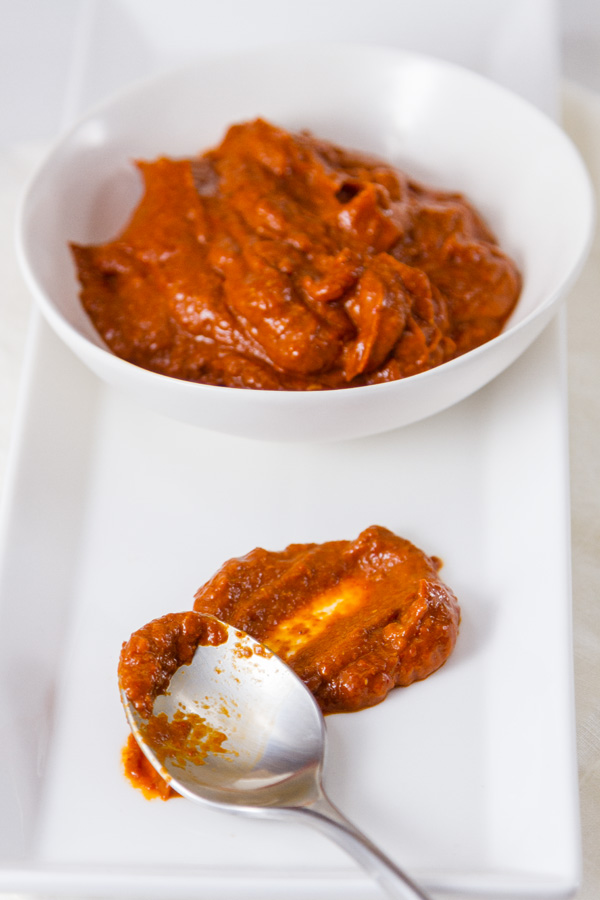a bowl filled with red spicy paste and a spoon in front of the bowl with a smear of the paste all on a white plate