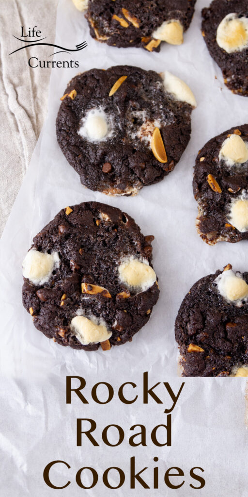 chocolate cookies topped with almonds and marshmallows on a piece of parchment paper, title on bottom: Rocky Road Cookies.