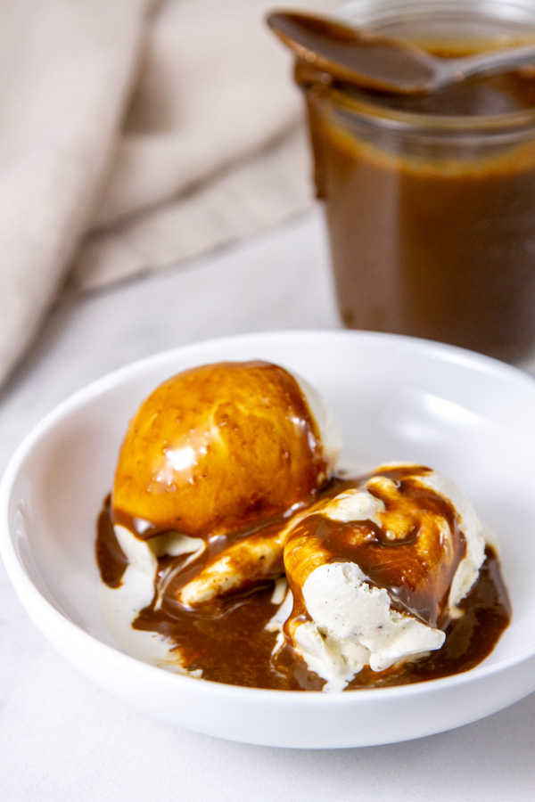 a white bowl with vanilla ice cream and sauce on top with the jar in the background.