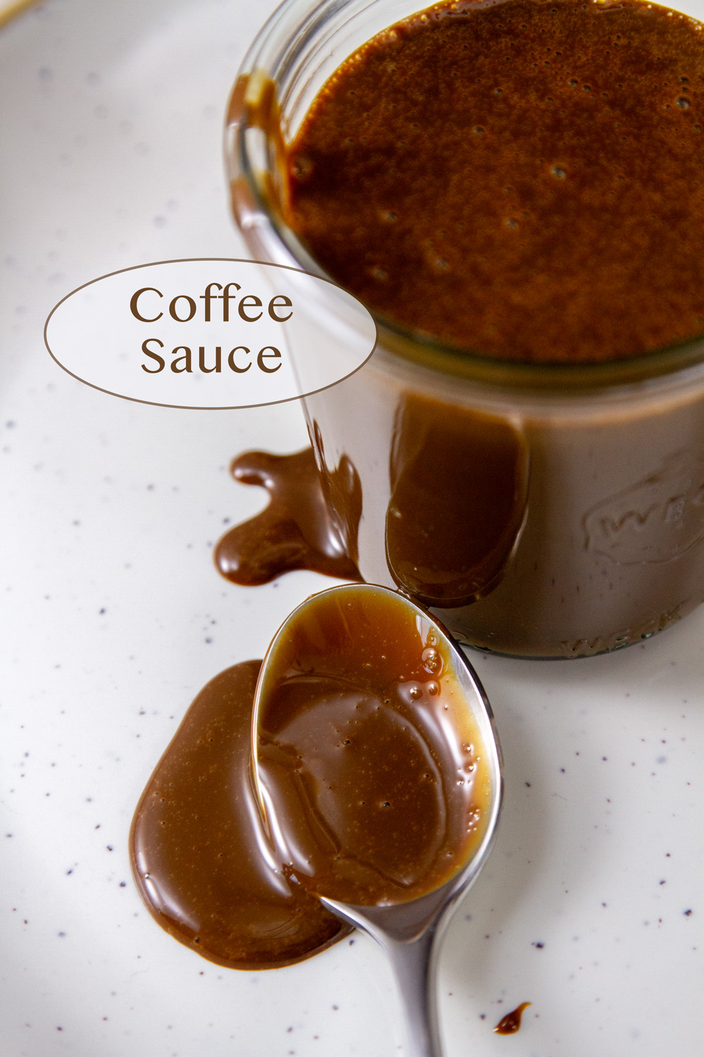 a glass jar with coffee sauce in it next to a spoon with the sauce flowing out of it.