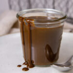 a glass jar filled with coffee sauce and some has spilled over the side, there's a spoon to the right.