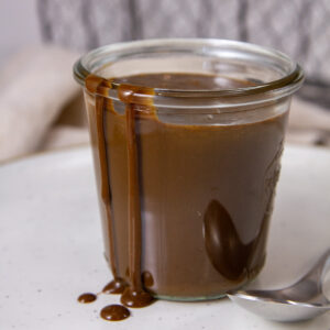 a glass jar filled with coffee sauce and some has spilled over the side, there's a spoon to the right.