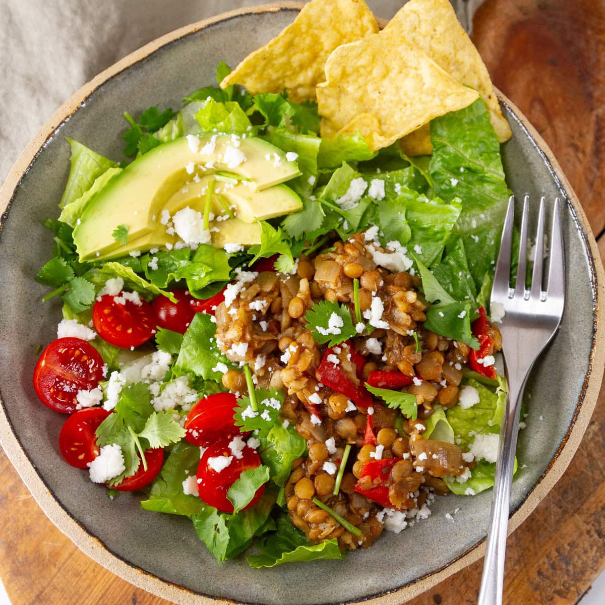 square crop of a bowl of salad with lentils, tomatoes, avocado, and chips, a fork on the right.
