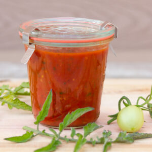 a glass canning jar filled with homemade marinara sauce and some tomato leaves and a small green tomato around the jar.