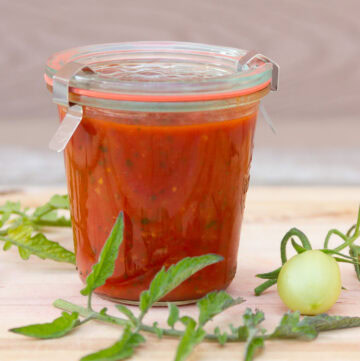 a glass canning jar filled with homemade marinara sauce and some tomato leaves and a small green tomato around the jar.