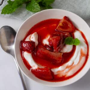square crop looking down on a bowl of yogurt topped with roasted strawberries and their juices with a spoon to the left and fresh mint around.