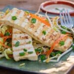 two slices of grilled tofu on cabbage slaw with a dish of peanut sauce in the background.