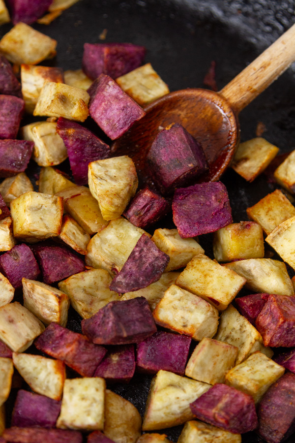 cubes purple and white sweet poatoes roasted in an iron skillet with a wooden spoon in the right.
