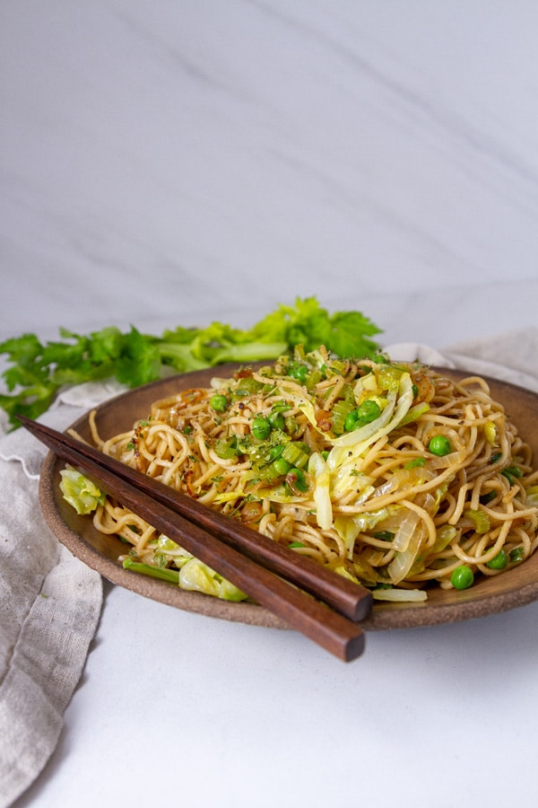 a plate filled with a cooked noodle dish and chopsticks on the side of the plate, a cloth napkin and celery next to the plate.