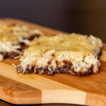 square crop of a brownie bar topped with coconut on a wooden cutting board.