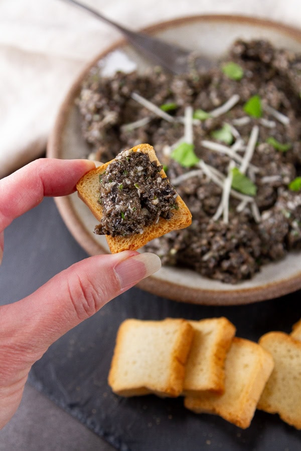 a hand holding a small toast with tapenade on it with the serving bowl in the background.