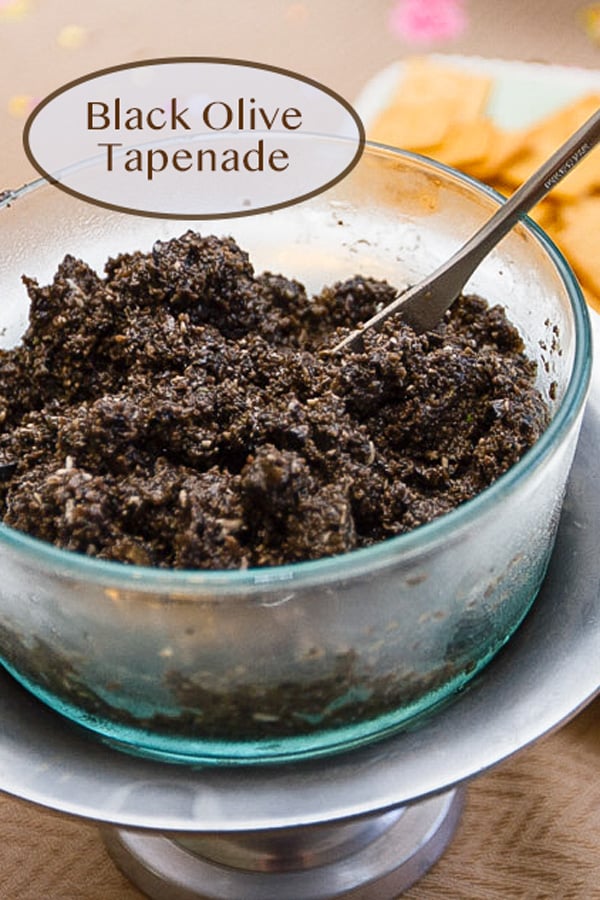 a glass bowl filled with black olive tapenade and a spoon sticking out of it.