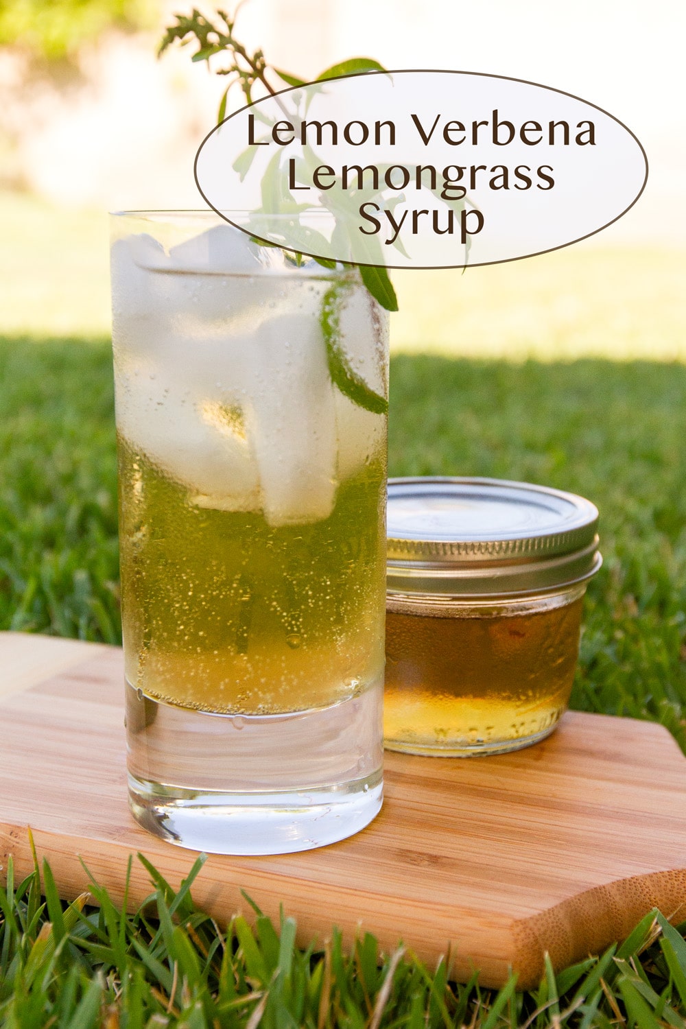 a glass filled with ice and a drink in front of a mason jar of syrup on a wooden cutting board on grass.
