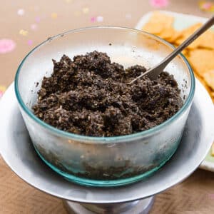 square crop of a glass bowl filled with tapenade and a spoon sticking out, crackers in the background.