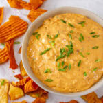 square crop of a bowl of dip made with sun dried tomatoes and garnished with green onions chips scattered around the bowl.