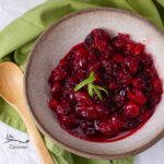looking down into a bowl filled with cranberry sauce, a wooden spoon and green napkin are next to the bowl.