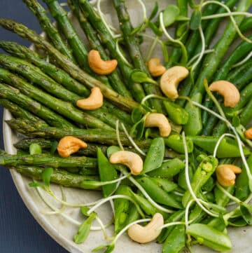 square crop looking down on a plate of spring vegetables asparagus, green beans, snap peas topped with sprouts and cashews.