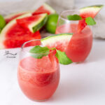 square crop of two glasses filled with red drink, watermelon slices behind.