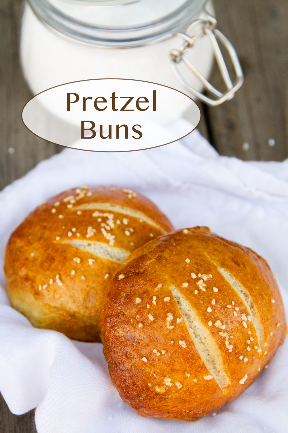 two pretzel buns on a white cloth napkin with a glass jar of pretzel salt in the background.