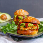 two sliders on pretzel buns on a dark plate with cilantro and an avocado.