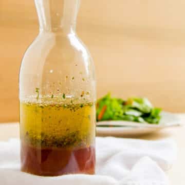 Square crop of a glass bottle filled with Italian Dressing in front of a salad.