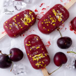square crop of several red Cherry Popsicles decorated with glitter and there are cherries and ice cubes next to them.