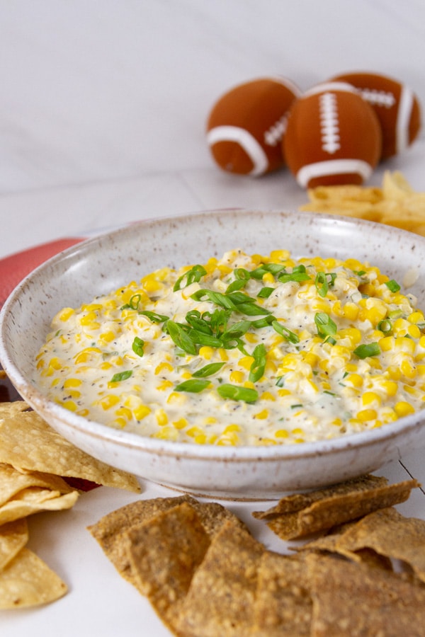 a bowl full of corn dip with chips and small footballs around it.