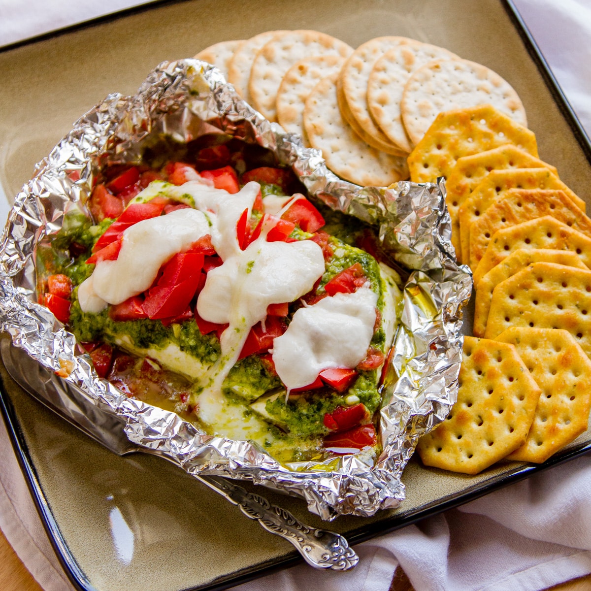 looking down into a foil packet filled with cheese, tomatgoes, pesto, served with crackers.