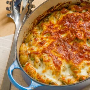 square crop of a cheesy topped casserole with noodles and mushrooms in a blue casserole dish and a serving spoon next to it.
