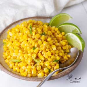 square crop of Caribbean Corn side dish in a bowl with lime wedges and a spoon.