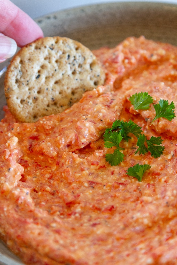 dipping a cracker into red pepper dip that's garnished with fresh parsley.