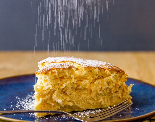 sprinkling powdered sugar on top of a slice of cheese cake with a fork next to it.