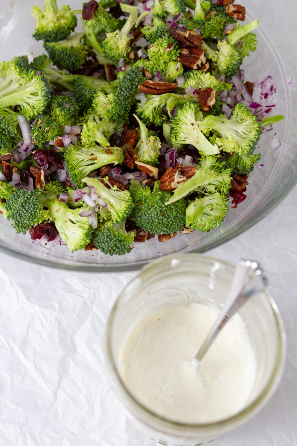 a large glass bowl filled with fresh broccoli florets, pecans, red onion, and cranberries next to a mason jar filled with creamy dressing and a spoon sticking out.