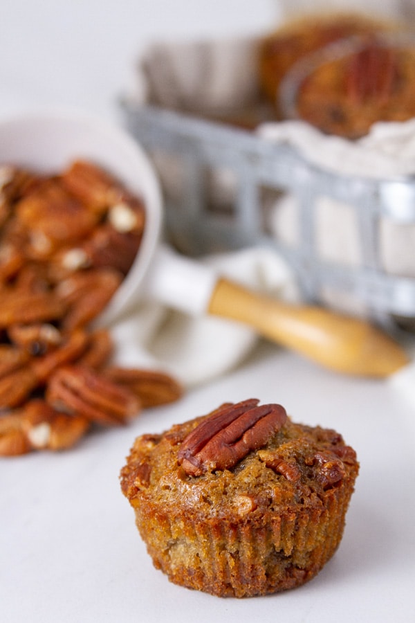 a muffin in front of a wire basket filled with more muffins and a measuring cup that's spilled out pecans.