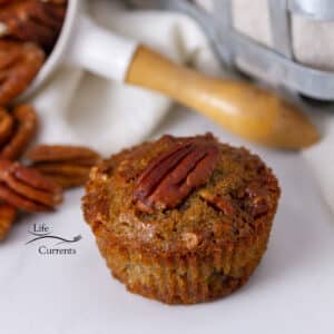 a single muffin with a pecan half on top in front of a measuring cup filled with pecans and a cloth napkin.