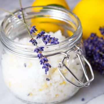 a glass jar filled with sugar scrub for your skin and topped with lavender flowers lemons and lavender behind the jar.
