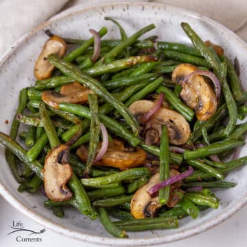 square crop of a bowl of cooked green beans with onions and mushrooms.