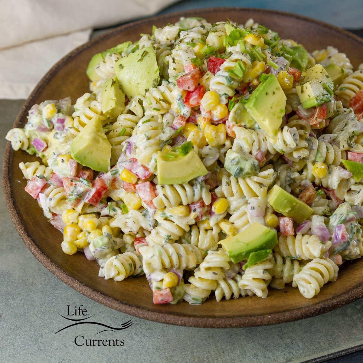 a brown bowl filled with pasta salad with avocado, red peppers, and corn.