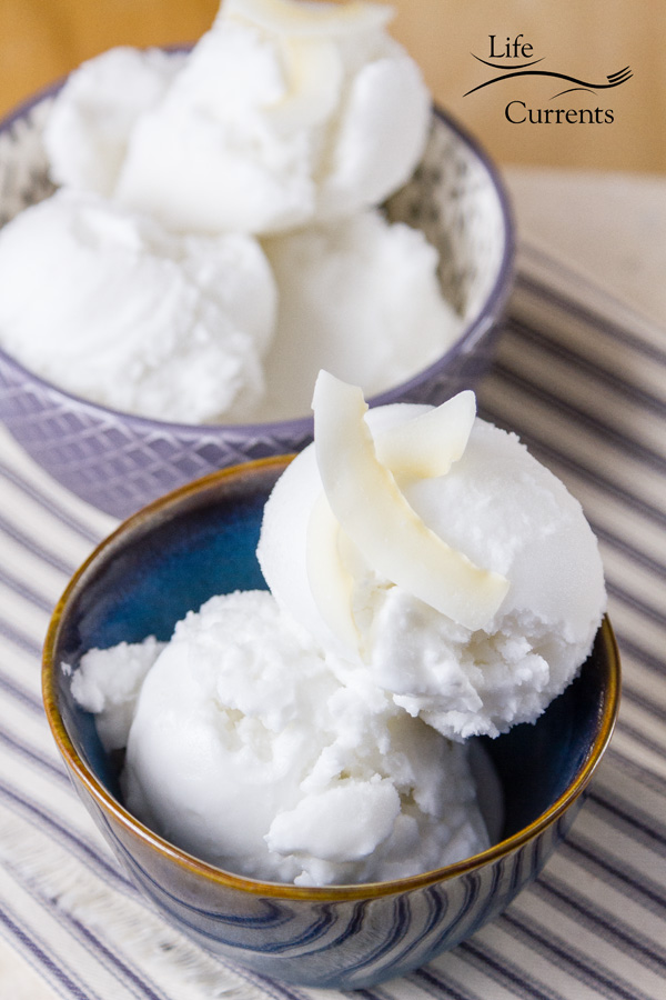 Two small bowls of coconut sorbet, the one in front is blue and the one in back is lavender colored. Both on a striped blue cloth