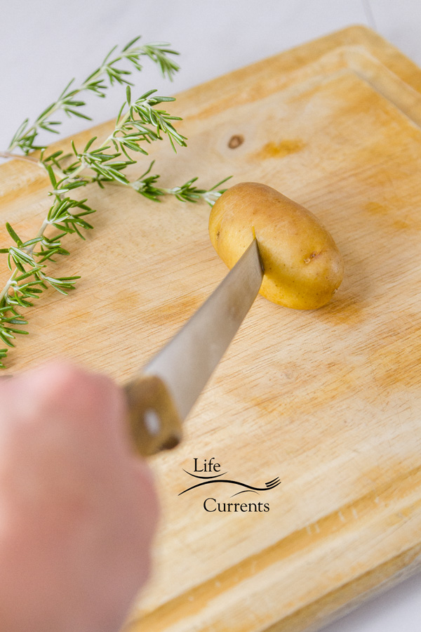 Piercing the potato with a sharp knife on a wooden cutting board with a sprig of fresh rosemary next to it 