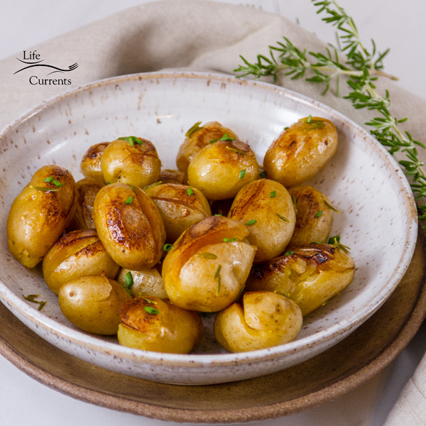 Cooked baby Yukon Gold potatoes with crispy browned edges in a white bowl on a brown bowl with fresh rosemary in the background