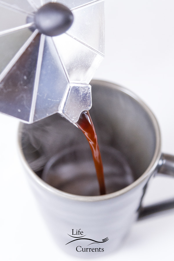 Coffee being poured from the stovetop espresso machine into a dark gray cup with steam coming off