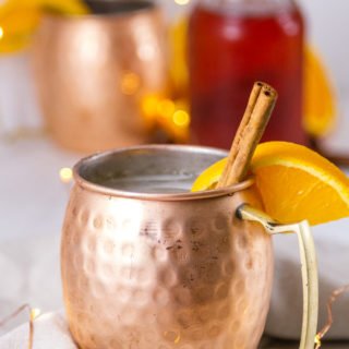 Copper mugs with orange and cinnamon garnishes with a bottle of cinnamon syrup in the background and twinkle lights around
